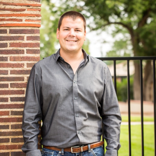Smiling man with short hair wearing a gray button-up shirt and blue jeans, standing outdoors next to a brick wall and iron fence with trees in the background.