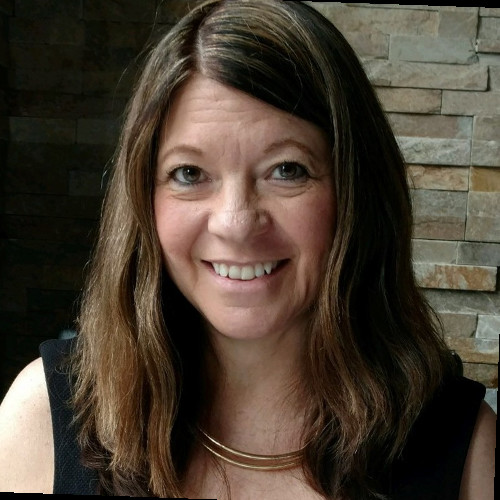 Smiling woman with long brown hair wearing a black sleeveless top and a gold necklace, posing in front of a stone wall.