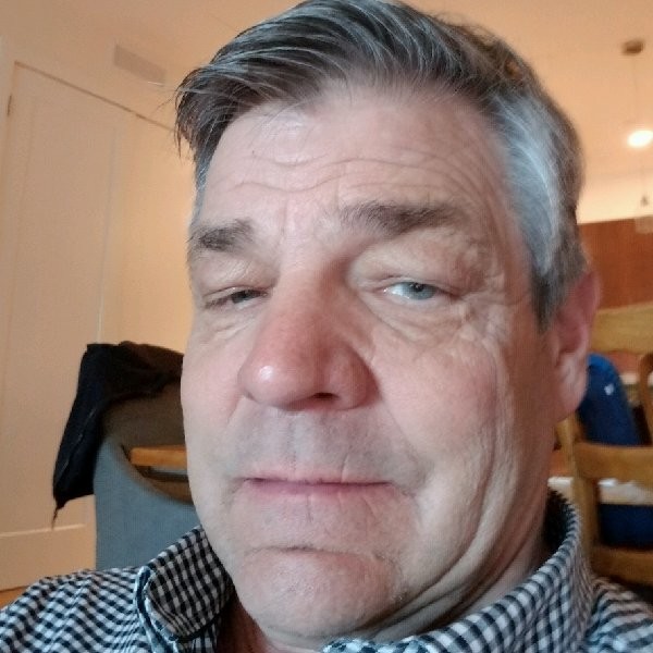 Close-up of an older man with gray hair, wearing a checkered shirt, sitting indoors with furniture in the background.