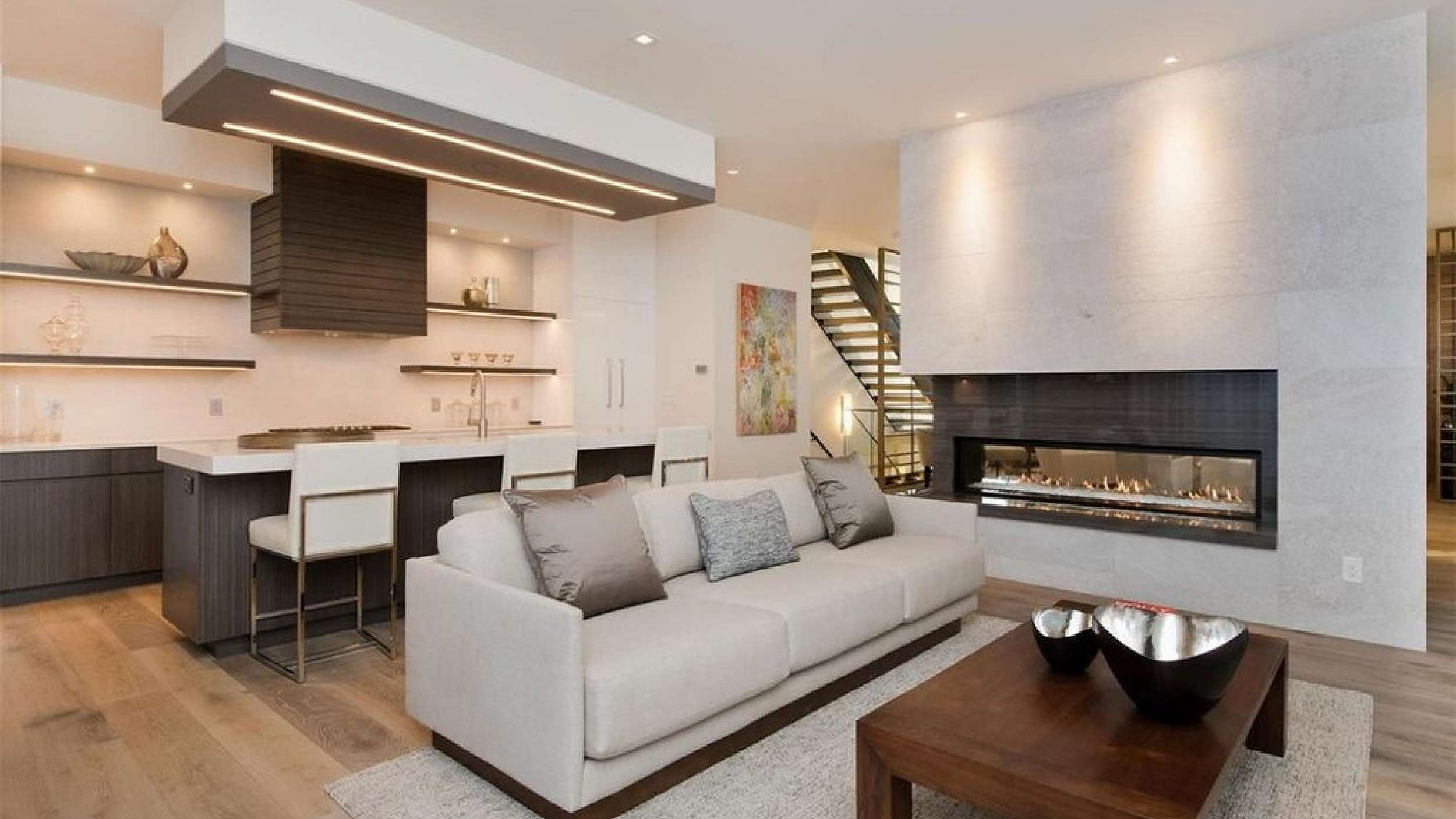 Modern open-concept living space with a light gray sofa adorned with three pillows, a wooden coffee table holding two decorative bowls, a built-in linear fireplace within a light stone wall, and an adjacent kitchen featuring a dark wood island with white bar stools, floating shelves with glassware, and a contemporary range hood.