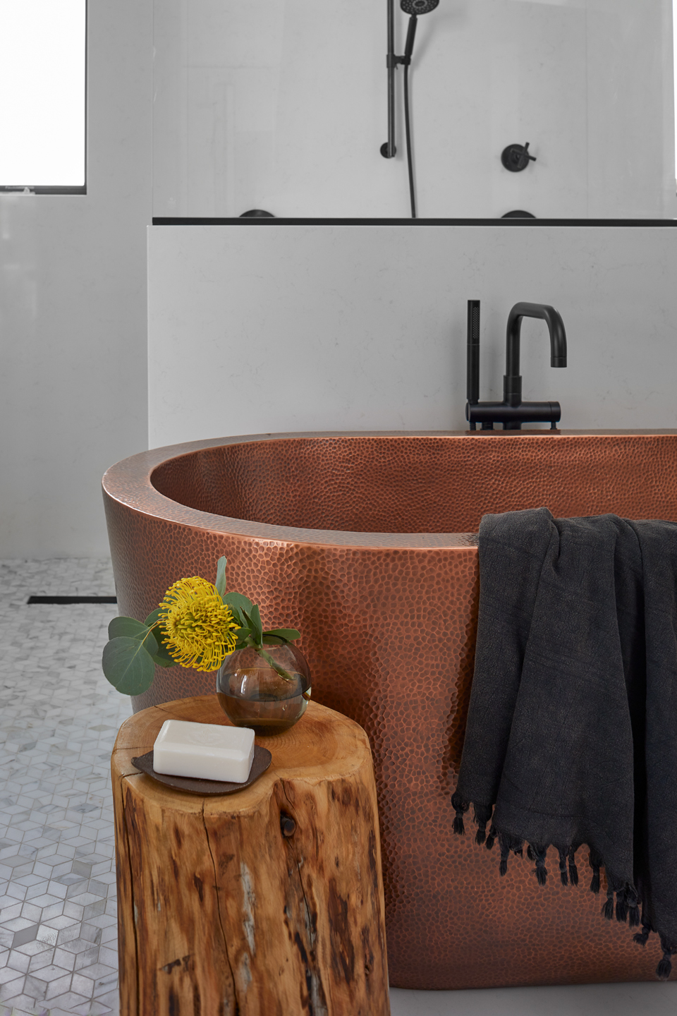 Copper textured bathtub with black faucet and black towel draped over the edge, next to a wooden stool holding a vase with a yellow flower and a bar of soap.