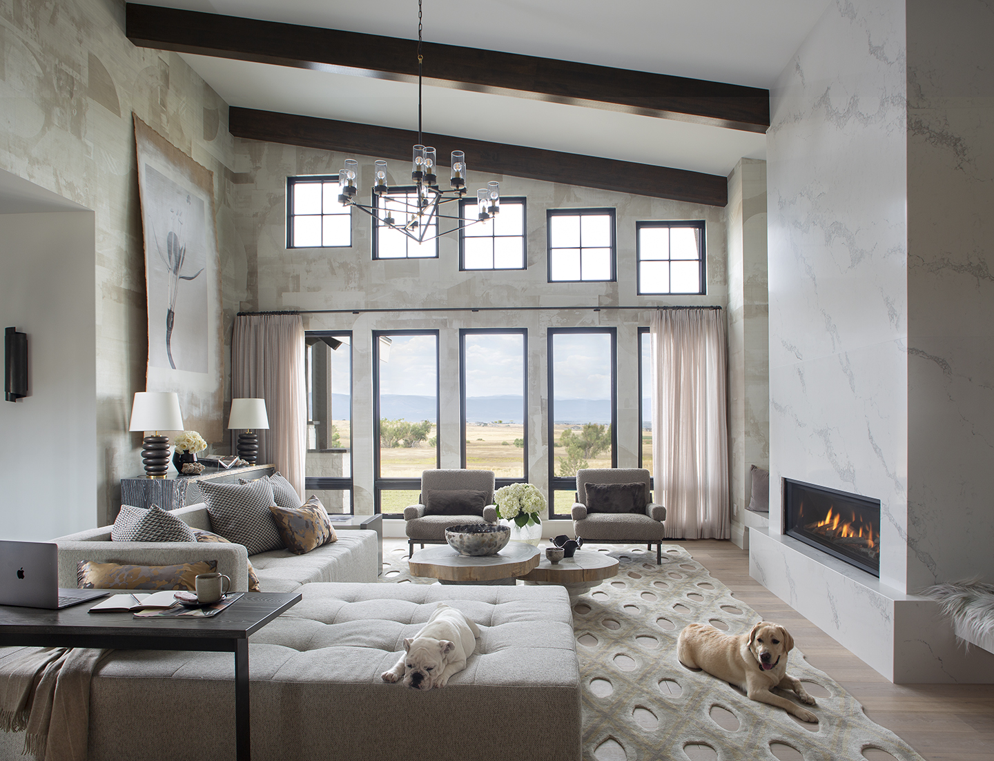 Modern living room with high ceilings and exposed dark wooden beams, featuring a large gray sectional sofa with patterned pillows, two gray armchairs, a set of round wooden coffee tables, and a fireplace with a marble surround on the right. A beige rug with large circular cutouts covers the floor. A white bulldog sleeps on the sofa chaise, and a yellow Labrador lies on the rug near the fireplace. Large windows with pale pink curtains offer a view of an open field and distant mountains under a cloudy sky. A dark wooden side table with a laptop and a coffee cup sits next to the sofa.