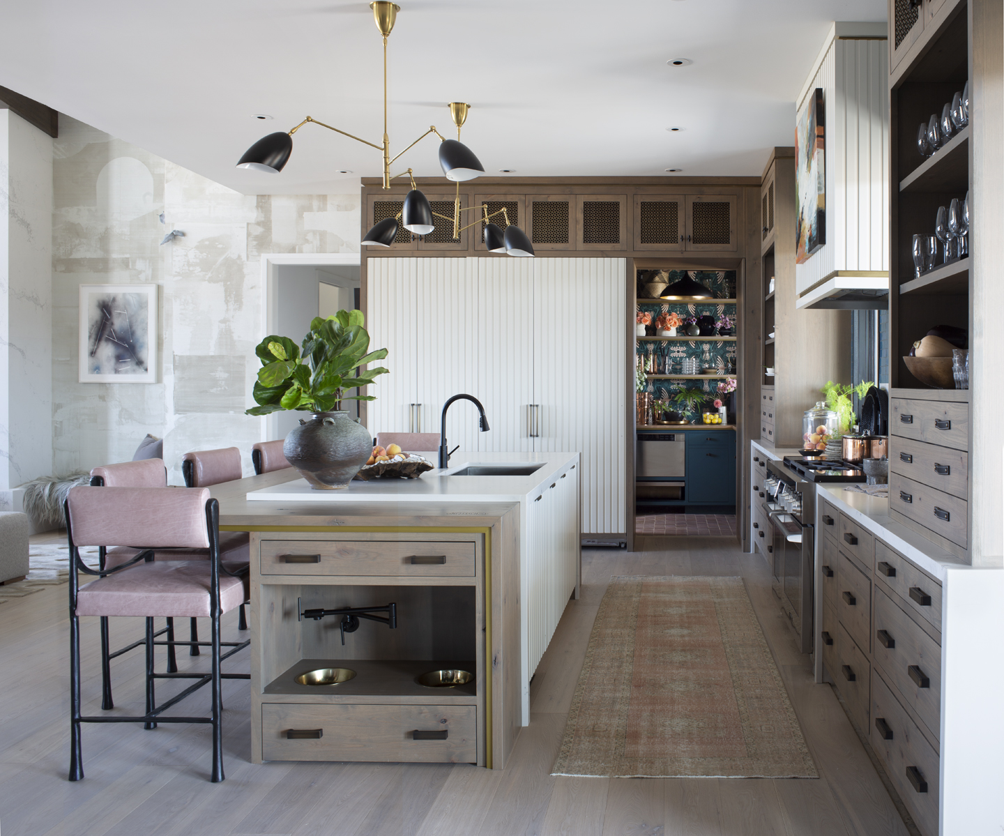 Modern kitchen with light wood cabinetry, a white island featuring a built-in pet feeding station, black faucet, and pink cushioned stools, illuminated by black-and-gold pendant lights.