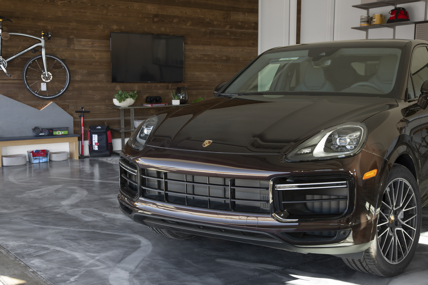 Dark brown Porsche SUV parked inside a modern garage with a concrete floor, wooden wall, mounted bicycle, TV, and shelves with various items in the background.