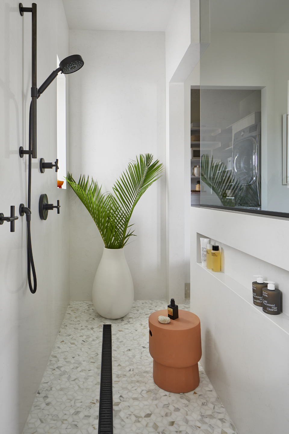 Modern minimalist shower area with white marble hexagonal tile floor, black shower fixtures, a large white vase with green palm leaves, an orange ceramic stool holding a bottle and soap, and built-in shelves in the wall with various bottles, reflected in a glass partition showing a washer and dryer in the adjacent room.