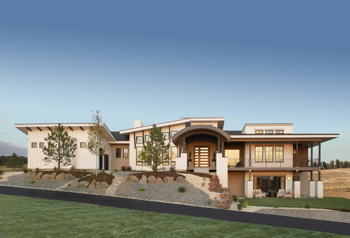 Modern two-story house with a mix of white siding and stone exterior, large windows, an arched wooden front door entryway with stairs, and a spacious covered balcony overlooking a landscaped yard with rocks, trees, and grass under a clear blue sky.