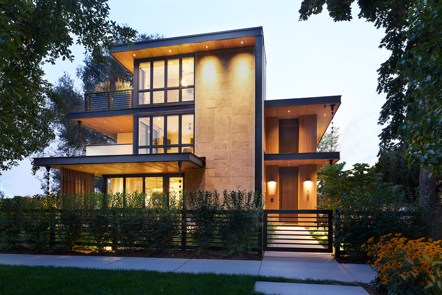 Modern three-story house at dusk with large windows, warm interior lights, vertical stone and wood cladding, a small front garden with bushes and a black gate, framed by trees and flowers.