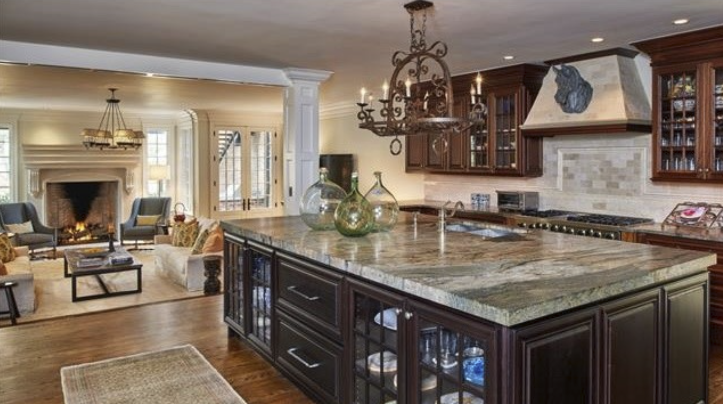 Kitchen with dark island and veined stone counter open to a family room with stone fireplace
