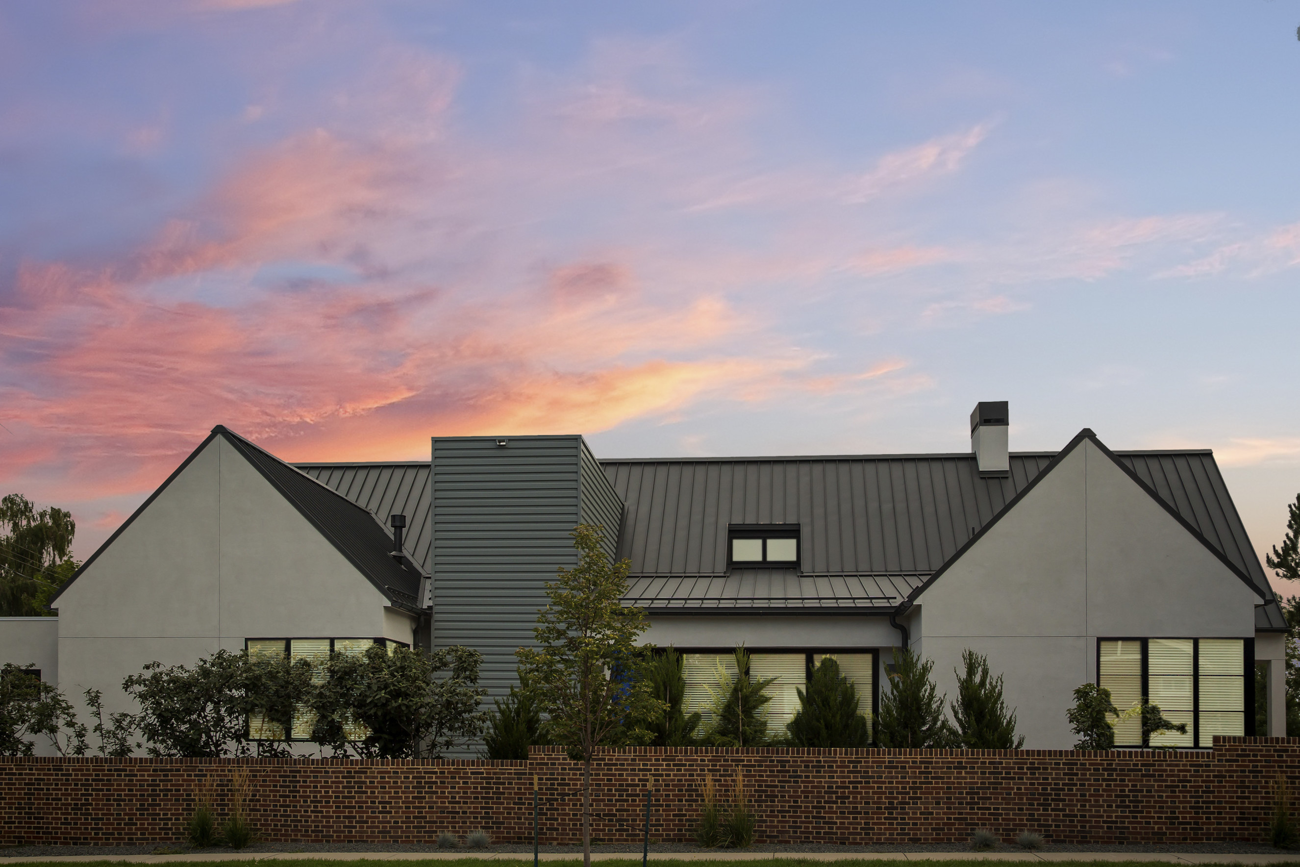 Modern farmhouse with standing-seam metal roof behind a brick garden wall at sunset
