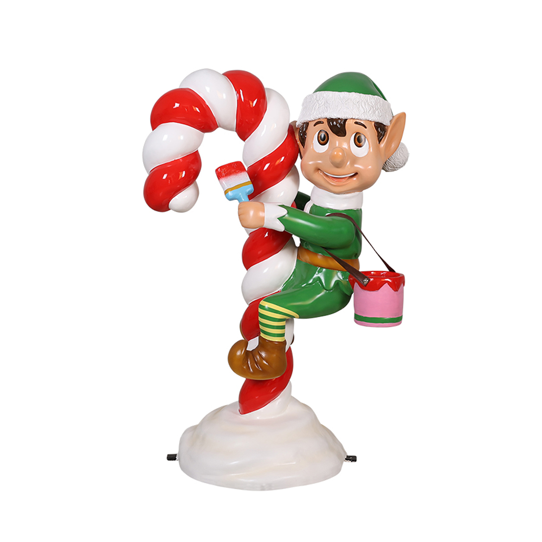 Christmas elf in green outfit hanging on and painting a large red and white candy cane with a paintbrush.