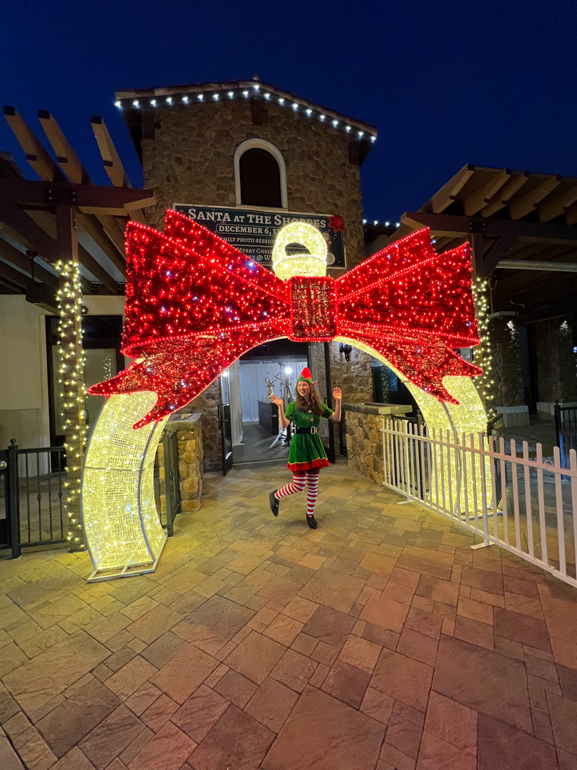 Person dressed as a Christmas elf posing under a large light up ornament arch with an illuminated red bow at night.