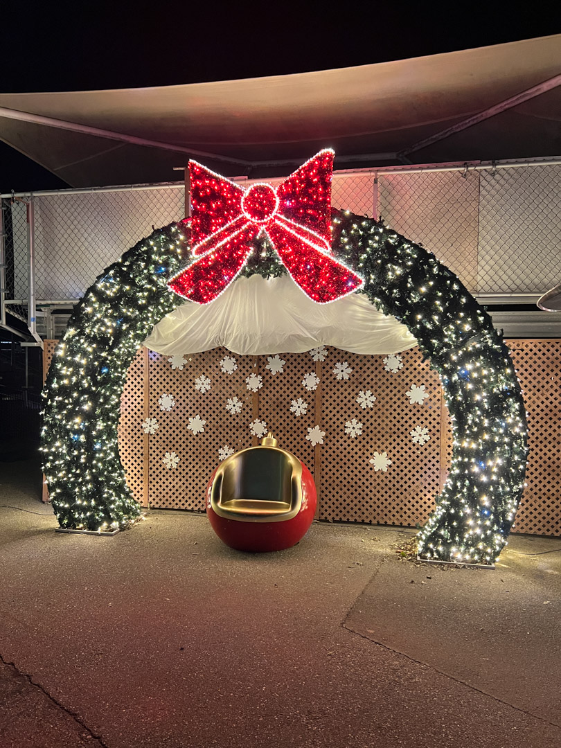 Christmas wreath arch with white lights and a large red bow above a red ornament-shaped chair.
