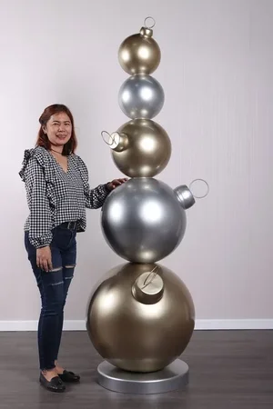 Woman standing beside a tall sculpture made of stacked metallic Christmas ornament balls in silver and gold.