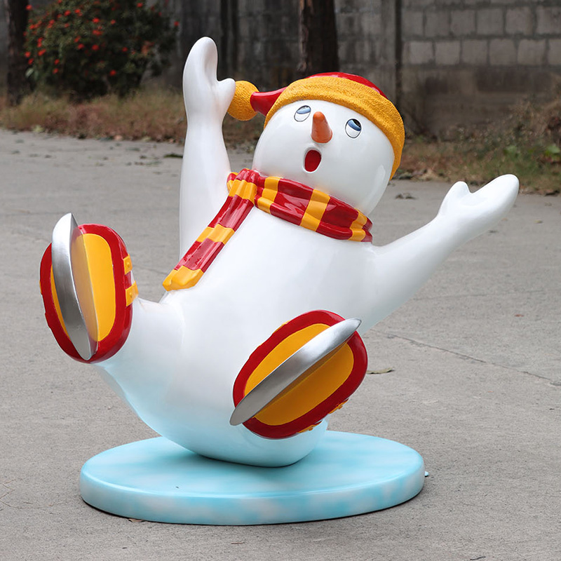 Playful snowman statue with orange hat, red and yellow scarf, slipping and sliding on blue base.