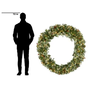 Green holiday wreath decorated with small lights and pine cones beside silhouette of a standing 72 inch tall man.