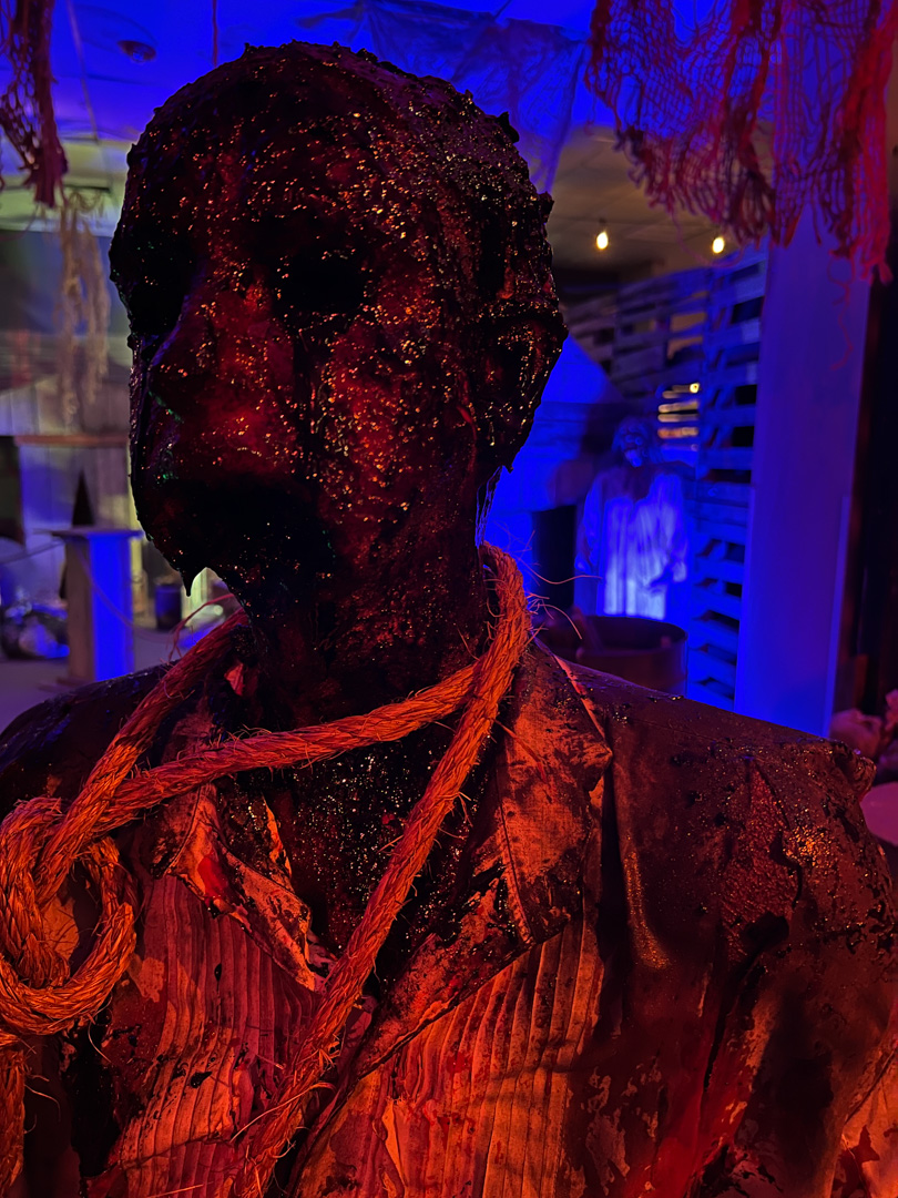 Dark, gruesome figure with a rope noose around its neck in a dimly lit, eerie room.