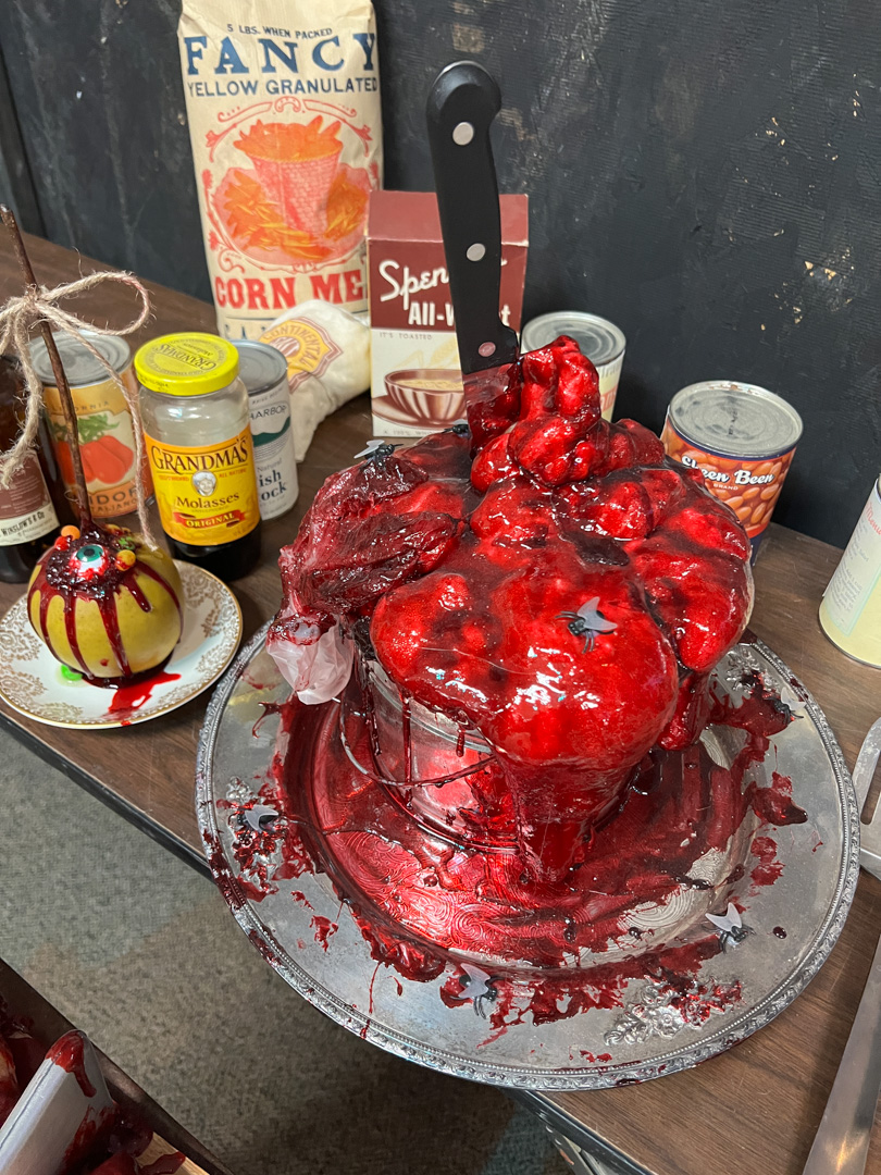 Red gelatinous cake made of organs with a knife stuck in top, fake flies on the surface, pantry items in background.