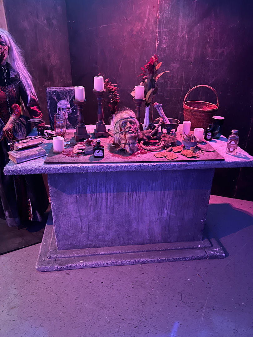 Table with Halloween props including a severed head, candles, bones, potions, and a figure in costume nearby.