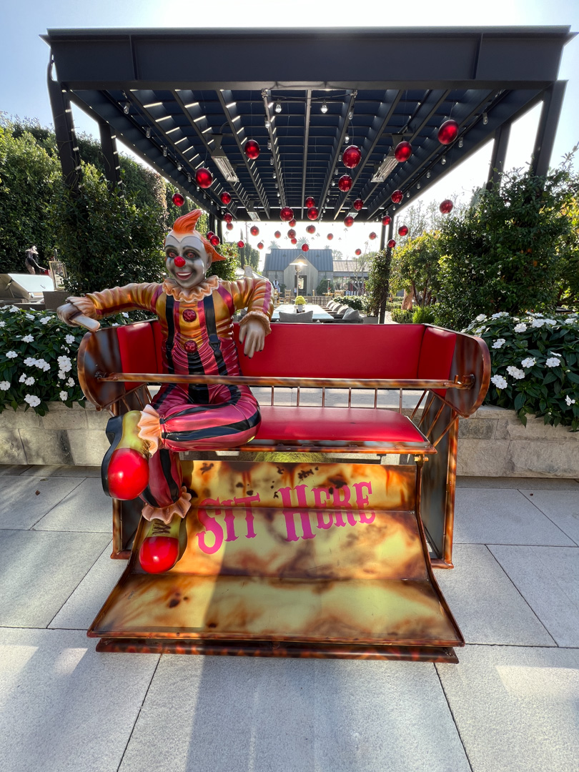 Colorful clown figure sitting on a red bench under a pergola with hanging red ornaments.