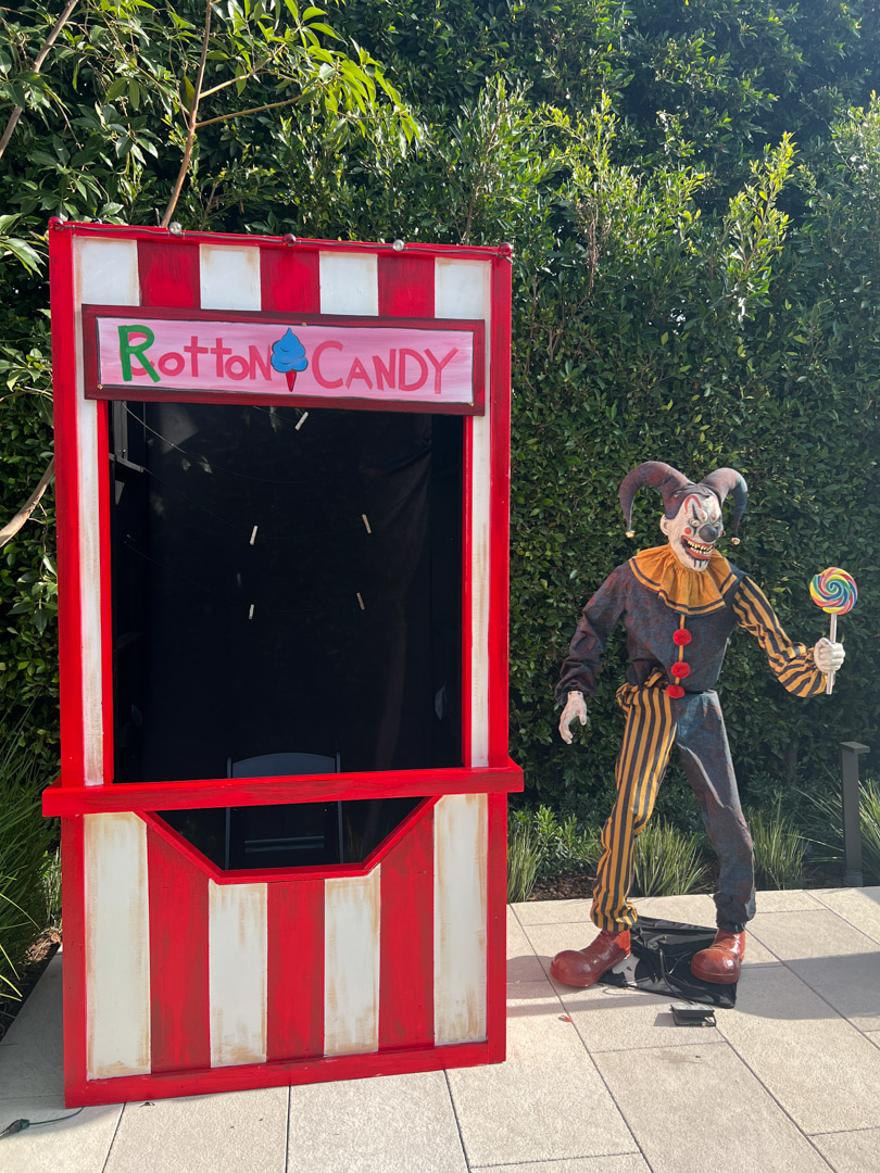 Red and white striped candy stand with sign 'Rotton Candy' next to a creepy clown holding a large lollipop.