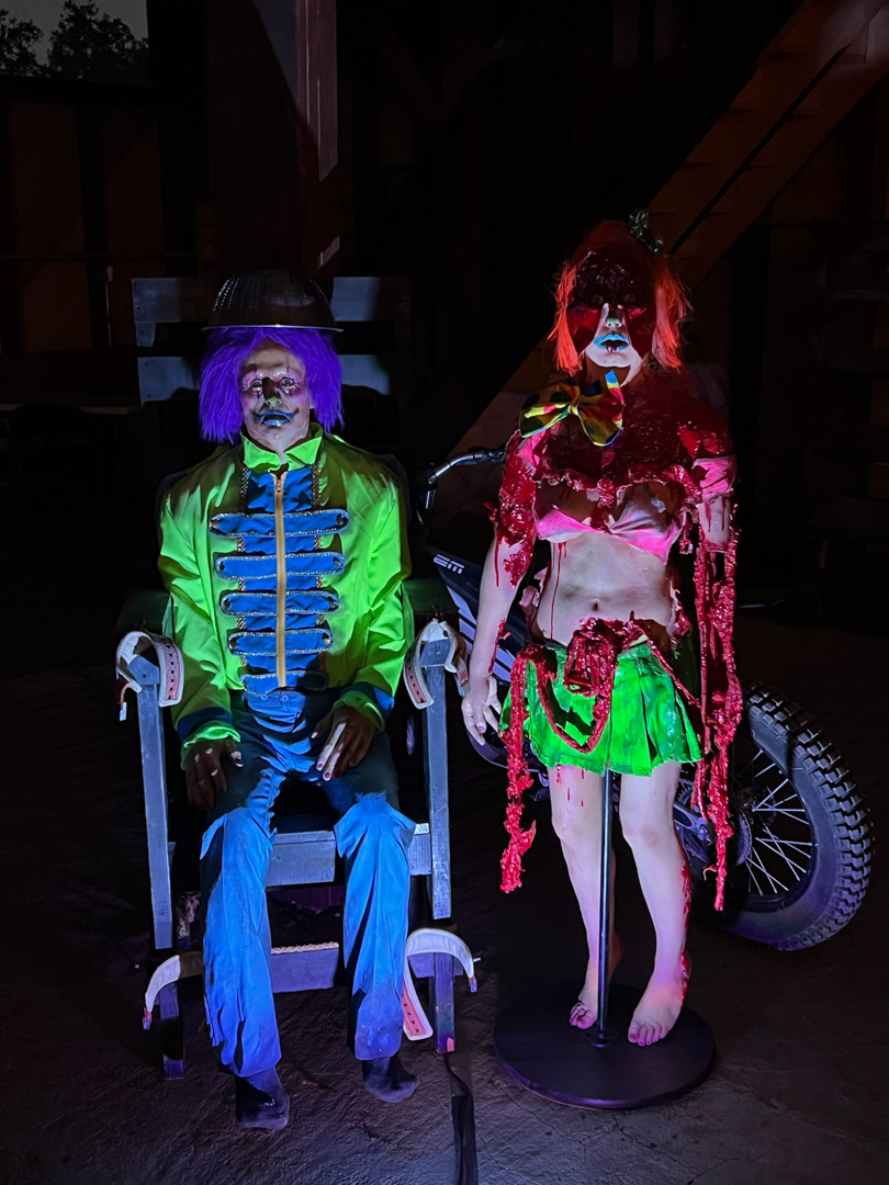 Two eerie clown figures in colorful costumes, one sitting and one standing, with dramatic lighting in a dark setting.