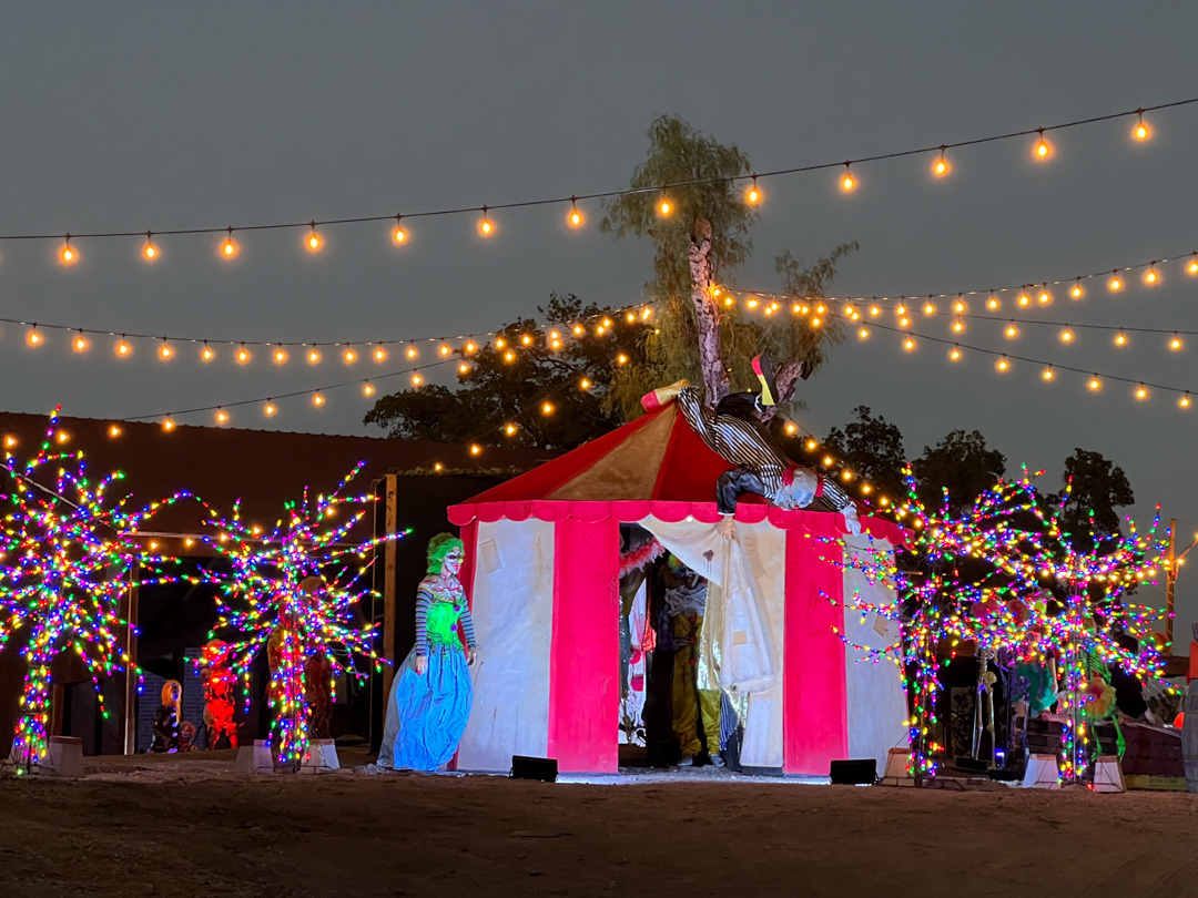 Night scene of a red-and-white circus tent with clowns and colorful light-decorated trees around it.