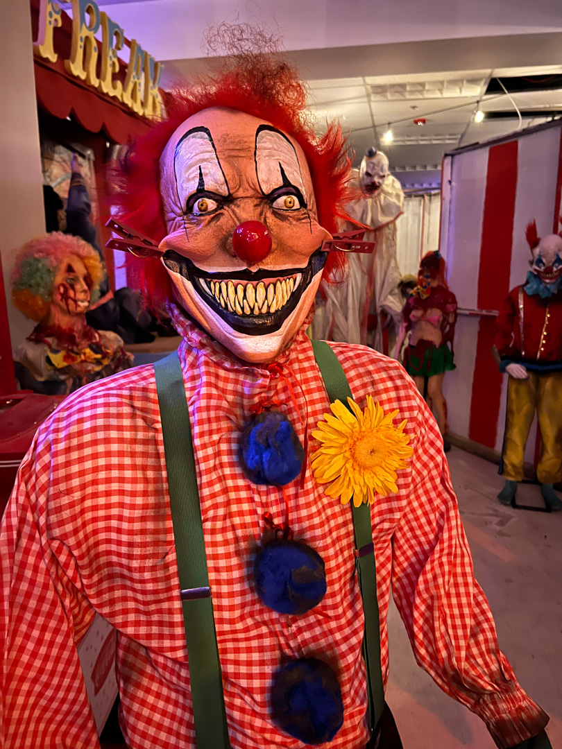 Scary clown mannequin with red hair, red nose, large teeth, red checkered shirt, and yellow flower.