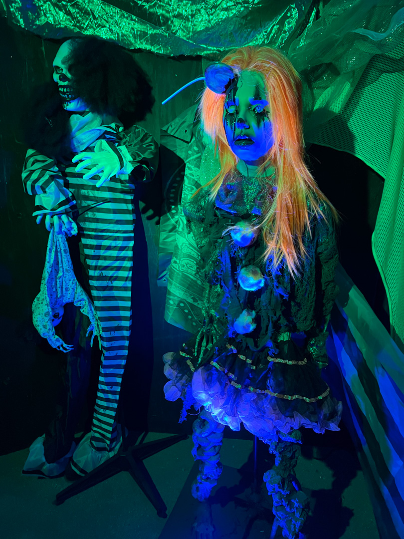 Two creepy Halloween figures under green and blue light: a skeleton clown and a doll with orange hair.