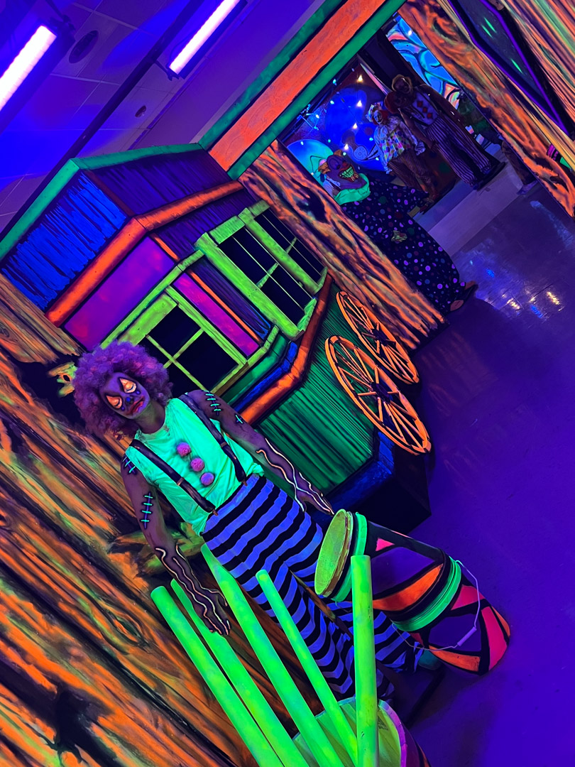 Three clowns with glowing makeup and costumes stand in a neon-lit maze with colorful props.