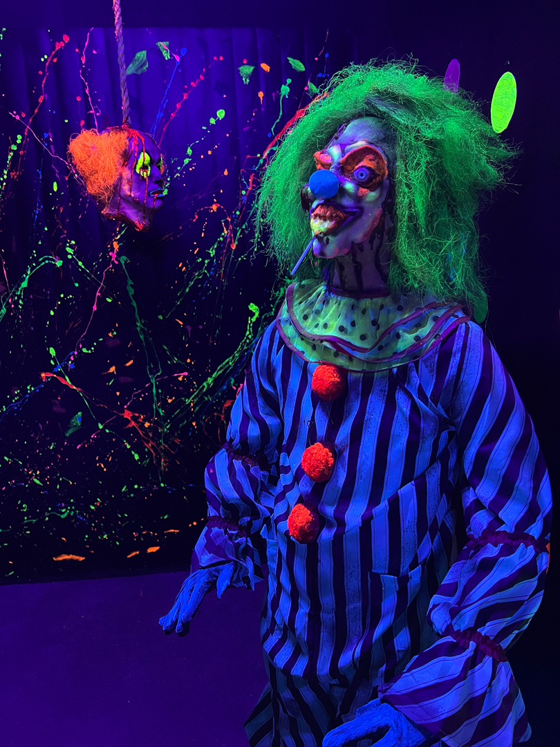 Scary clown with green hair, striped costume, and a lollipop with a severed clown head in the background.