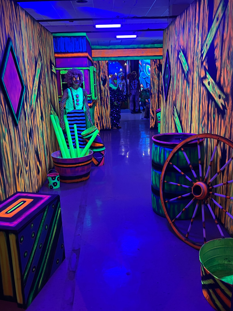 Neon-lit carnival hallway with colorful clown figures, props, and painted wooden walls.