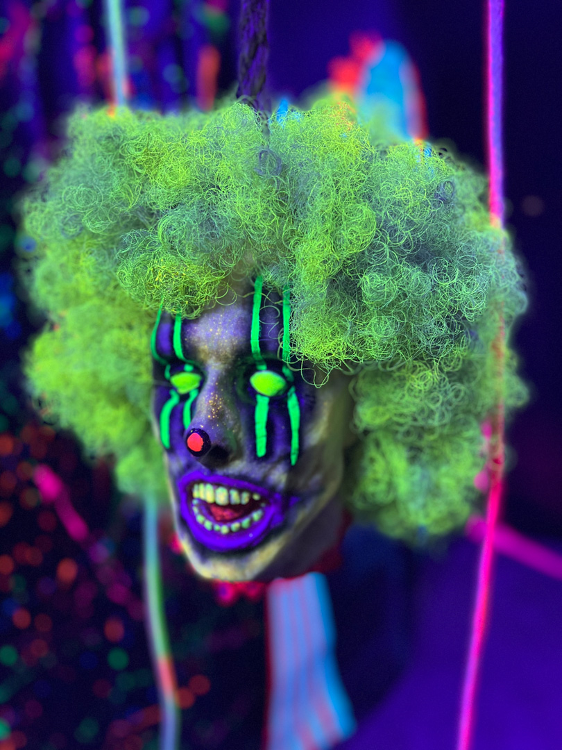 Glowing green-haired clown head with neon face paint and bright teeth in dark setting.