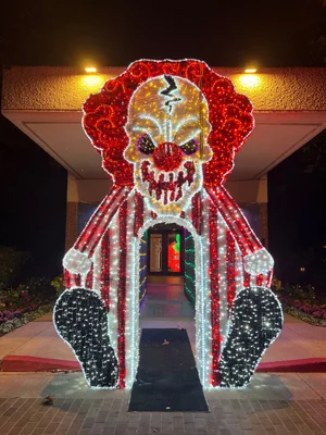 Lighted clown arch with red hair, striped suit, and big black shoes at night near a building entrance.