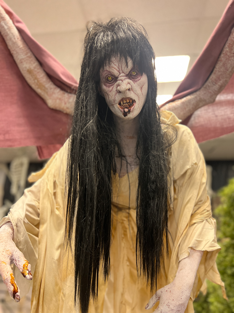 Scary creature with yellow eyes, dirty teeth, long black hair, pale skin, and tattered yellow dress with wings.