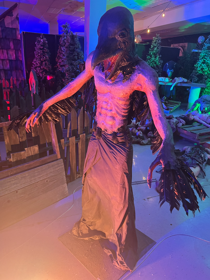 Scary humanoid creature with a bird-like head and wings standing in a dim, spooky room.