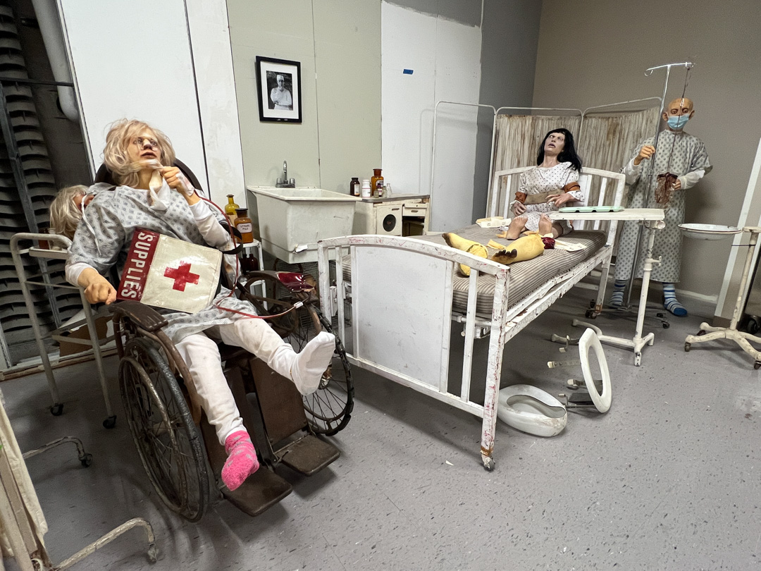 Hospital room with mannequins in gowns on wheelchair and bed, one standing with IV drip.