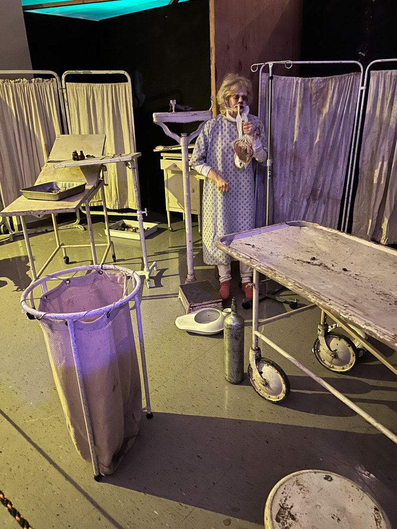 Woman in hospital gown holding a plastic bag around her neck in a room with old medical equipment.