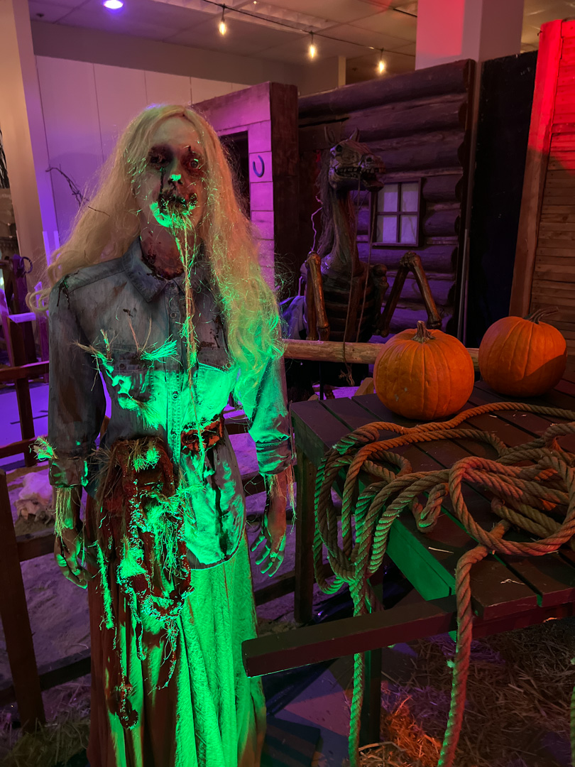 A creepy female figure stands among pumpkins under green light.