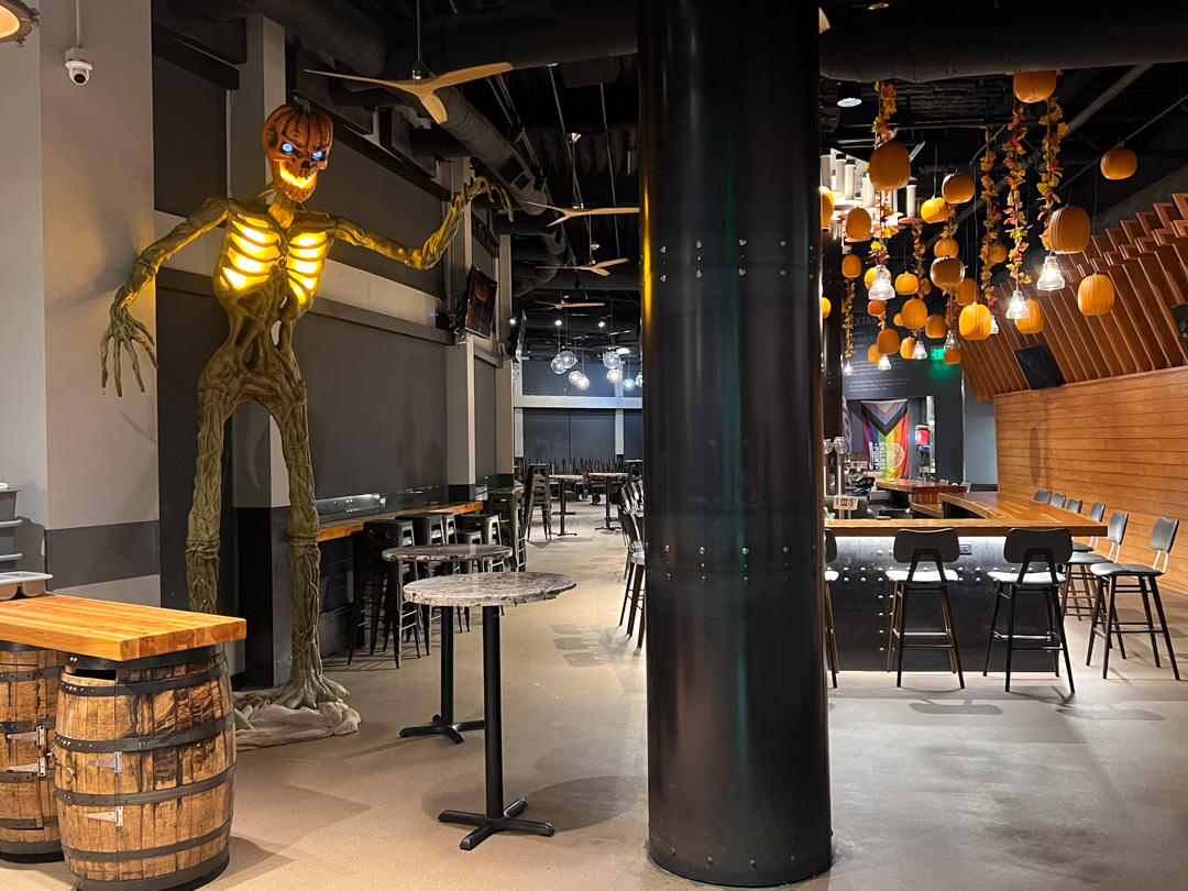A towering skeleton clings to a black support column beneath hanging pumpkins.