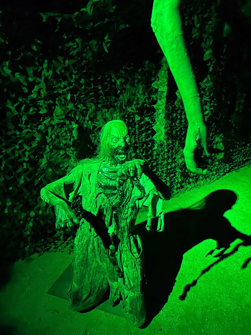 Green-lit zombie figure reaching out with a dismembered arm hanging above it casting a shadow.
