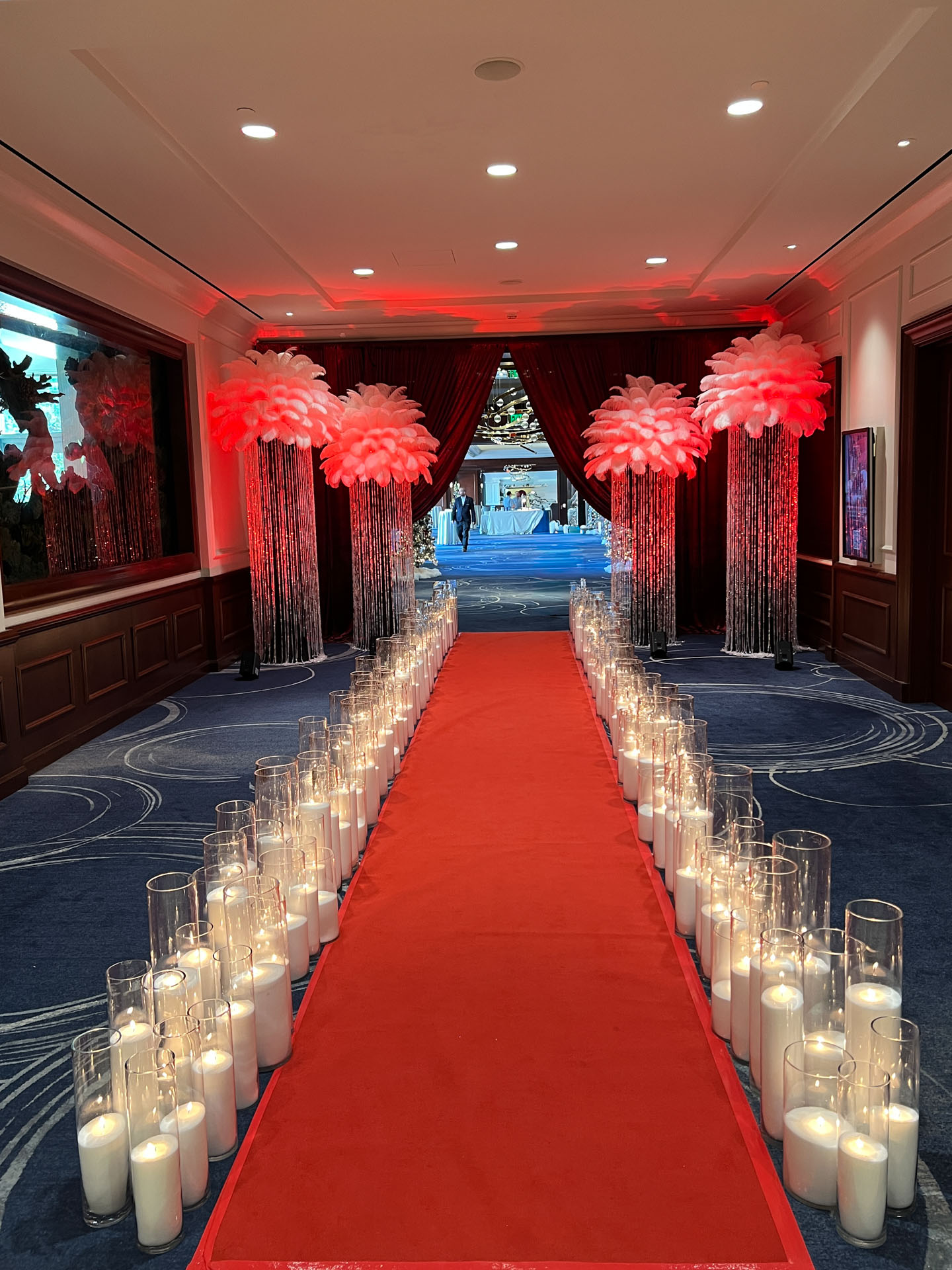 Red carpet aisle lined with glass candles and decorated with red-lit feather displays in an event venue hallway.