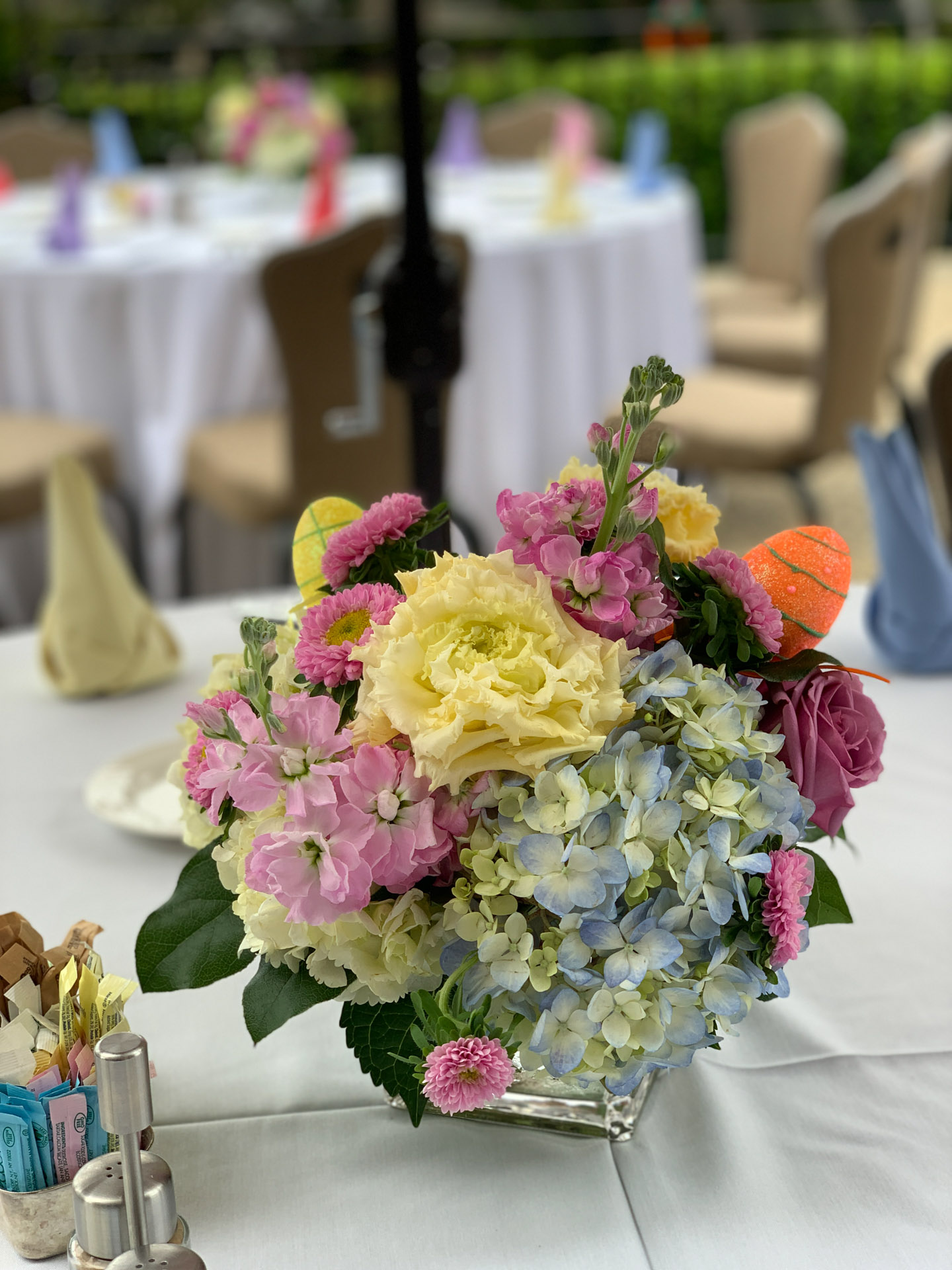 Colorful floral centerpiece with yellow, pink, blue flowers in glass vase on white tablecloth.