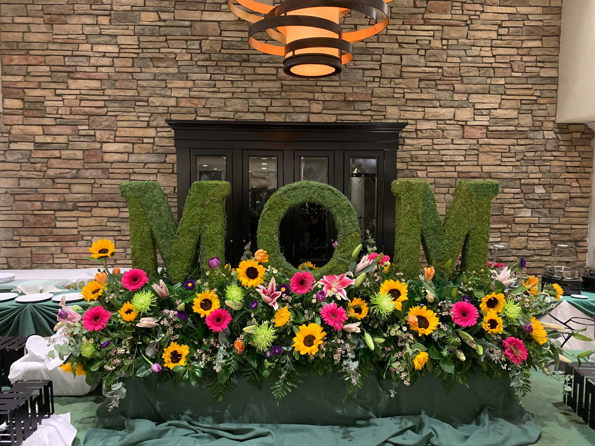 Green foliage letters spelling MOM surrounded by colorful flowers on a table with green cloth.
