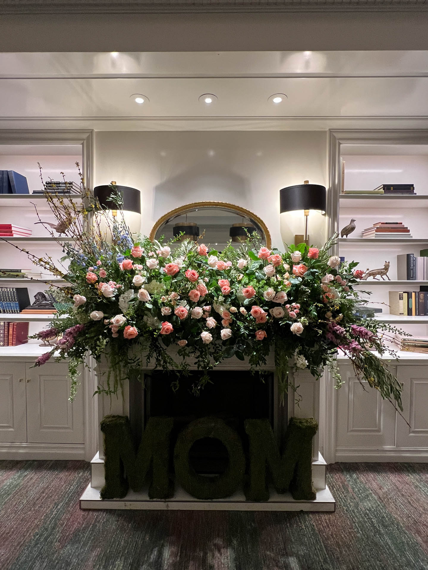 Fireplace decorated with a large floral arrangement of pink roses and greenery and foliage letters spelling MOM.