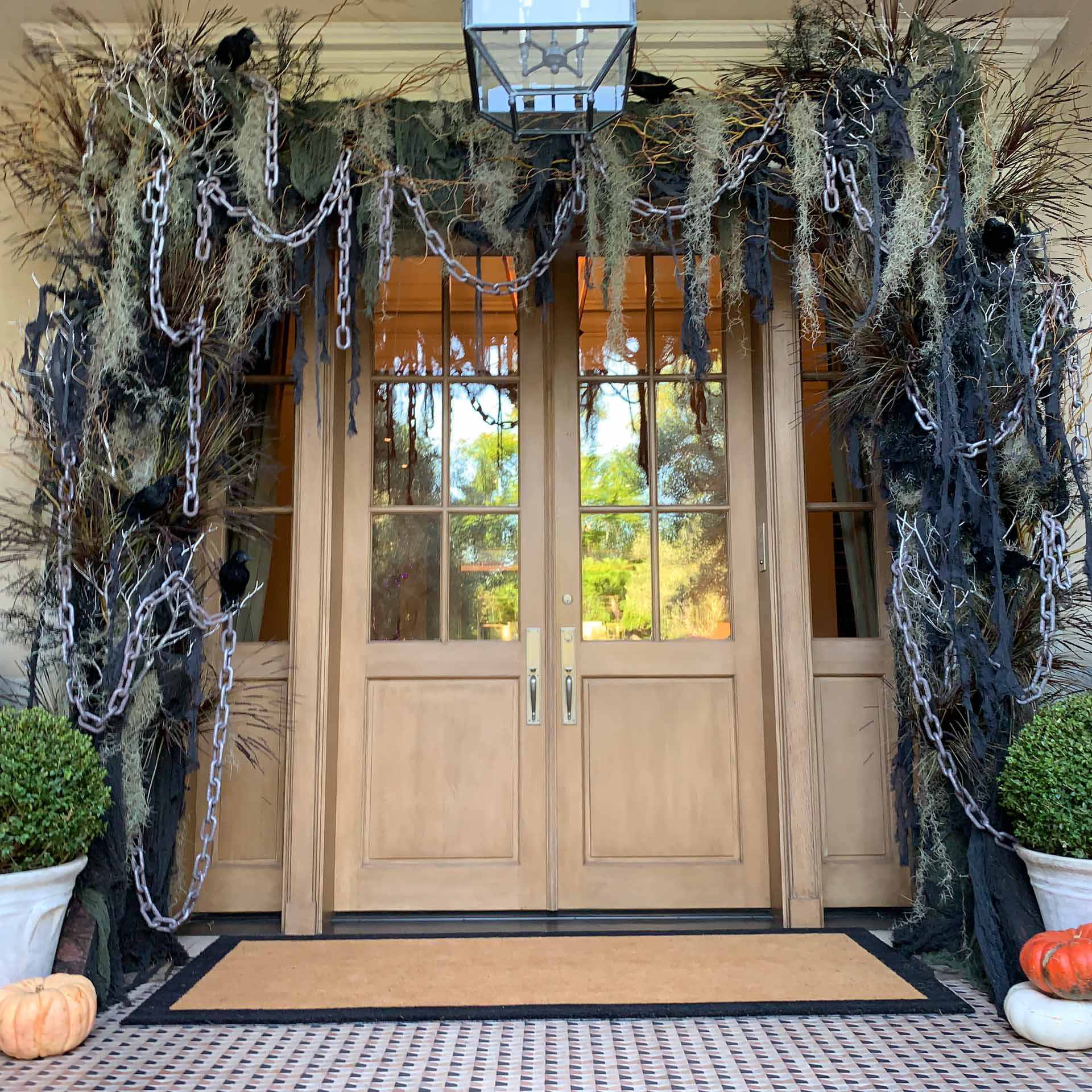 Front wooden double doors with Halloween decor: black chains, cobwebs, crows, and pumpkins on tiled porch.