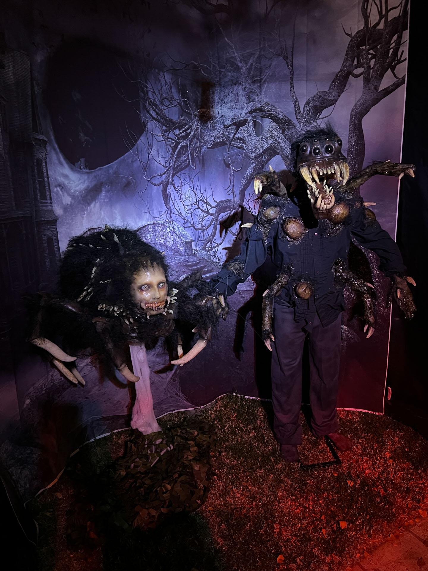 Two creepy spider creatures with human-like faces in a dark, eerie setting with a twisted tree backdrop.