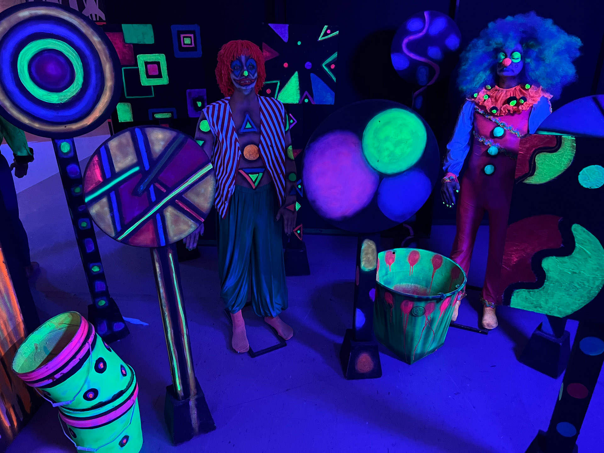 Two scary clowns in colorful costumes and makeup surrounded by neon abstract shapes under black light.