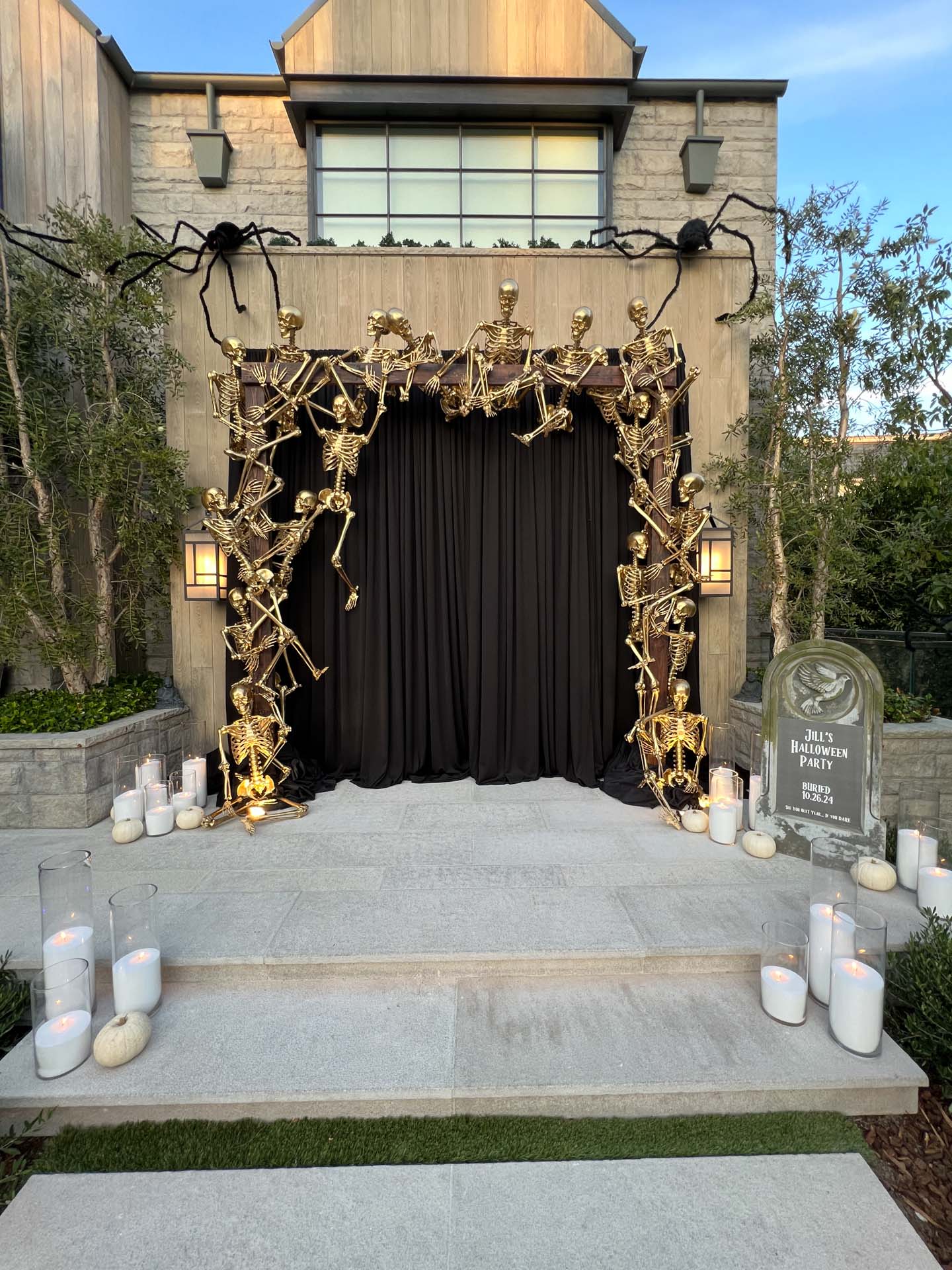 Gold skeletons decorate an archway with black curtains, candles, white pumpkins, and a tombstone sign for a Halloween party.