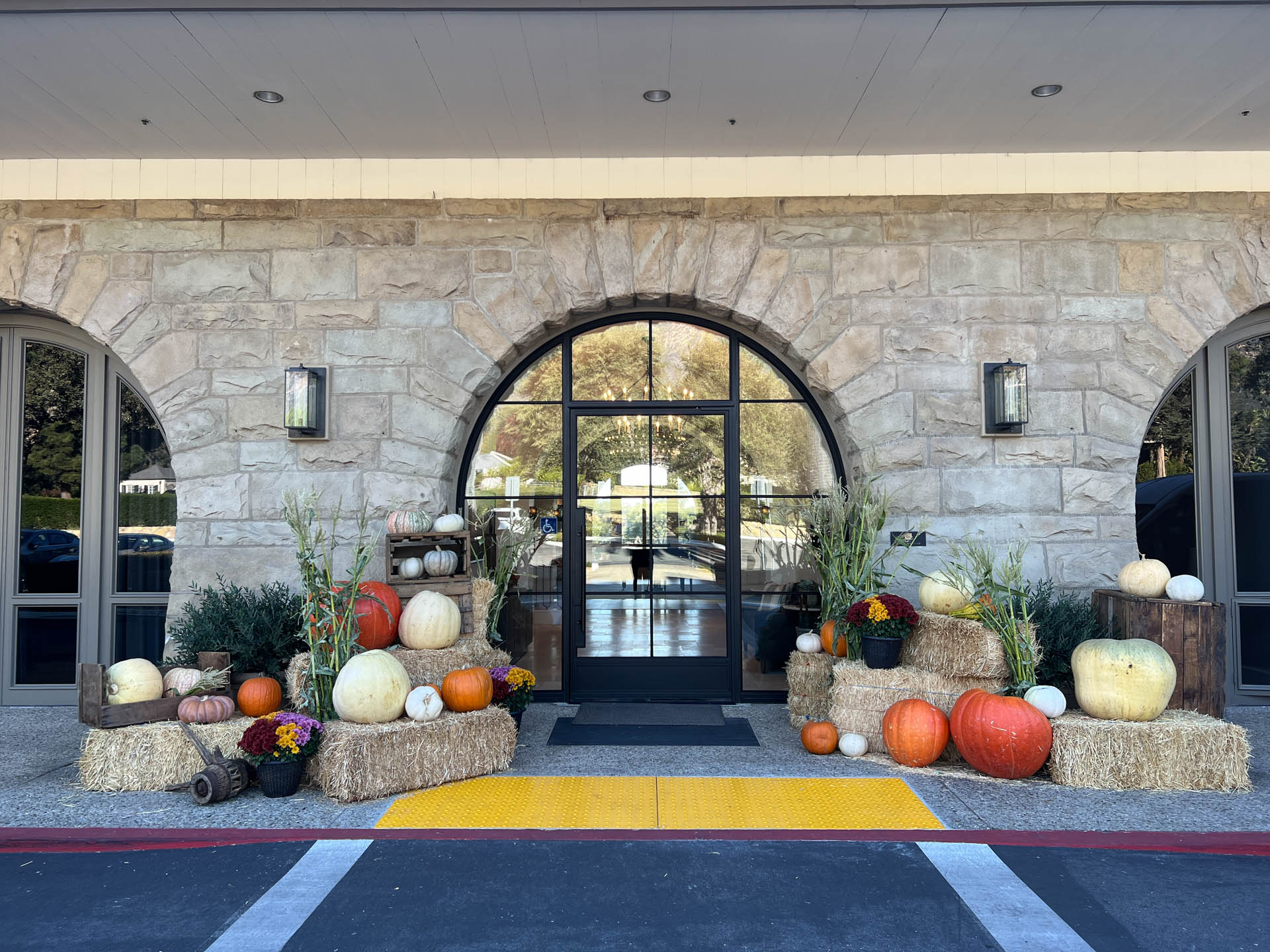 Thanksgiving decor with pumpkins, gourds, flowers, and hay bales arranged outside a stone building entrance.