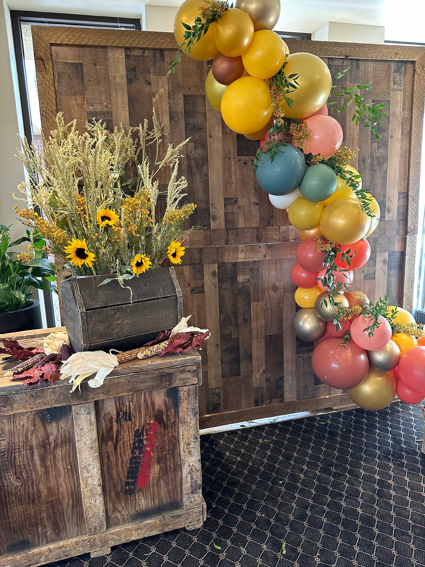 Rustic wooden crate with sunflowers and autumn leaves beside a colorful balloon arch with greenery.